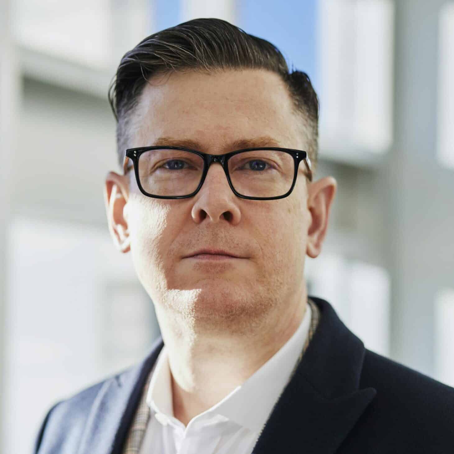 Profile portrait of a confident business executive wearing eyeglasses in a sunlit office setting.
