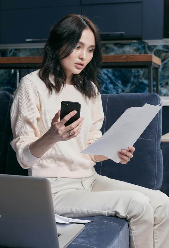 A young woman uses her smartphone and laptop to manage finances, she is shocked by the price of her Phones Bill.