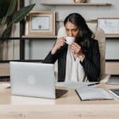 Woman sipping coffee while working on a laptop in a modern office.